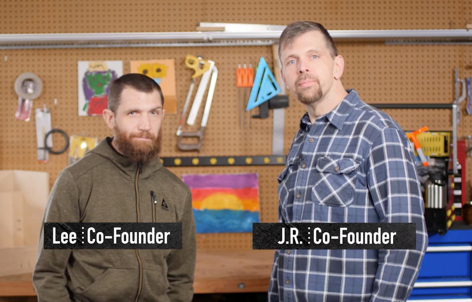 Trucker Tote founders: Lee and J.R. Photograph of Trucker Tote founders, Lee and J.R., who are friendly-looking, middle-aged white men wearing flannel shirts. They stand in front of a wall adorned with assorted workshop tools.
